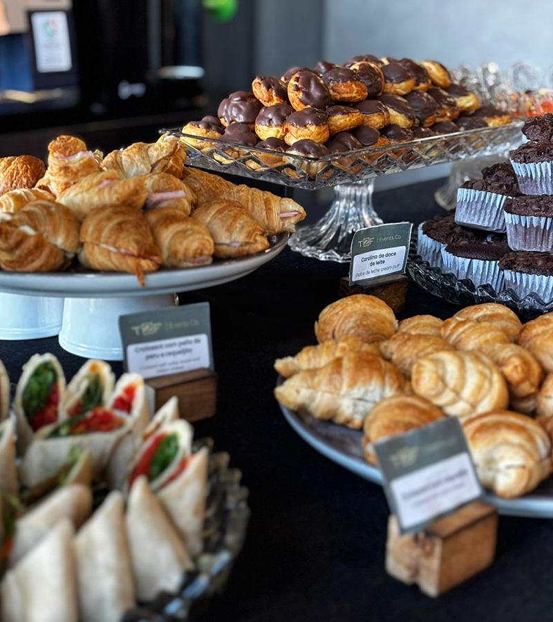 Mesa de coffee break corporativo com croissants, sanduíches, doces e salgados organizada para eventos empresariais